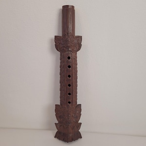 Op de afbeelding: Een donkerbruine houten fluit met ingewikkelde houtsnijwerk en zes gaten. De fluit is versierd met een gesneden gezicht aan de boven- en onderkant.