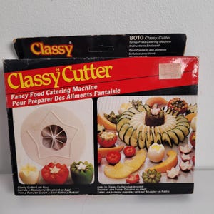 Op de afbeelding: Een Classy Cutter-doos met de productnaam in grote gele letters. De doos bevat afbeeldingen van creatief gesneden groenten en fruit. De doos bevat ook tekst in het Engels en Frans, waarin de mogelijkheden van het product worden aangeprezen.