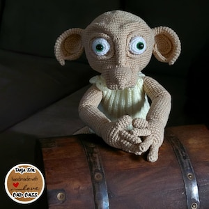 Könnte beinhalten: Eine gehäkelte Dobby-Puppe in einem gelben Kleid, die auf einem Holzfass sitzt. Die Puppe hat große grüne Augen und eine spitze Nase. Die Puppe ist handgefertigt mit Liebe.