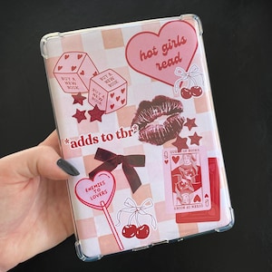 Puede incluir: Una funda protectora transparente para lector de libros electrónicos con un patrón de cuadros rosas y blancos. La funda está decorada con varias pegatinas, incluyendo dados, corazones, labios, cerezas, estrellas y naipes. El texto incluye "hot girls read".