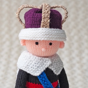 May include: A knitted doll wearing a purple and white crown, a white scarf, and a blue and red sash.