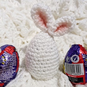 May include: White crocheted bunny rabbit egg cozy with pink ears. The bunny is sitting on a white fluffy surface next to two chocolate eggs wrapped in foil.