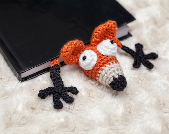 May include: A crocheted fox bookmark with orange and white fur, black eyes, and black legs. The bookmark is hanging off the edge of a black book.