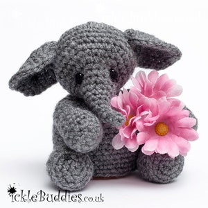May include: A gray crocheted elephant toy holding pink flowers. The elephant has large ears and small black eyes. The text "TickleBuddies.co.uk" is in the lower left corner. The toy is sitting on a white surface.