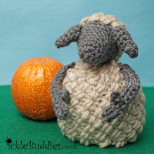 May include: A crocheted grey and white sheep toy with a fluffy white body and grey face. The sheep is sitting on a green surface next to an orange.