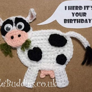May include: A crocheted white cow with black spots, pink nose, and googly eyes. The cow is holding green yarn in its mouth. A speech bubble above the cow says "I herd it's your birthday!"