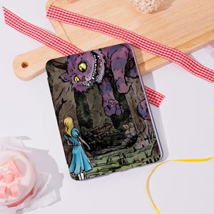 May include: A rectangular e-reader cover featuring a colourful illustration of a blonde figure in a blue dress standing before a large, grinning purple cat with yellow eyes in a forest setting, with a small sign labelled Tea Party visible.