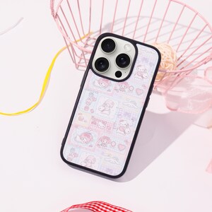 Op de afbeelding: Een zwarte telefoonhoes met een kleurrijk patroon met stripfiguren, harten en wolken. De hoes heeft een zwarte rand en een uitsparing voor de camera. De achtergrond bevat een roze draadmand en decoratieve linten.
