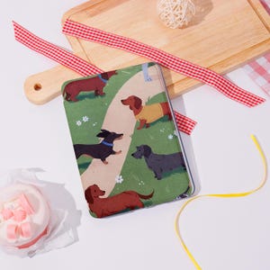 May include: A notebook featuring a whimsical illustration of dachshund dogs on a green background with a winding path. The dogs are in various colors, including brown, black, and gray. A red and white checkered ribbon and a wooden cutting board are in the background.