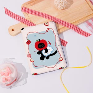 Puede incluir: Una funda blanca para tableta con un gato negro de dibujos animados con cabeza de manzana roja, sosteniendo una almohada azul. La funda tiene un fondo azul claro con detalles de manzanas rojas y estrellas. Una cinta a cuadros roja y blanca y una tabla de cortar de madera están en el fondo.