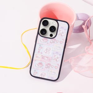 Op de afbeelding: Een zwarte telefoonhoes met een kleurrijk patroon met stripfiguren, harten en wolken. De hoes ligt op een wit oppervlak naast een roze mok en een roze decoratie-artikel.