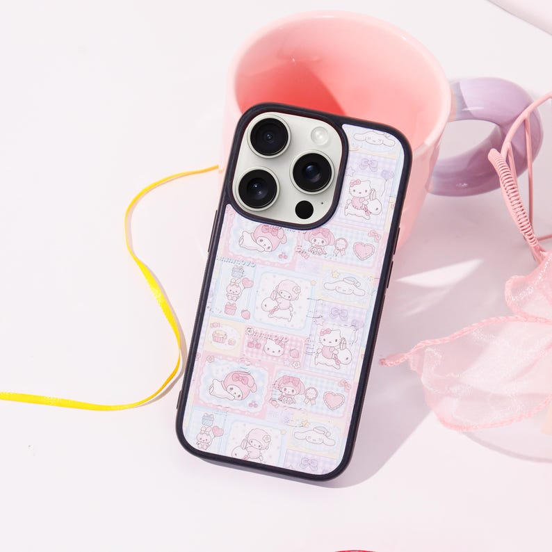 Op de afbeelding: Een zwarte telefoonhoes met een kleurrijk patroon met stripfiguren, harten en wolken. De hoes ligt op een wit oppervlak naast een roze mok en een roze decoratie-artikel.