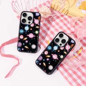 May include: Two black phone cases with a colourful space-themed design. The cases feature planets, stars, and comets in shades of pink, blue, orange, and purple against a black background. The cases are on a pink and white checkered surface.