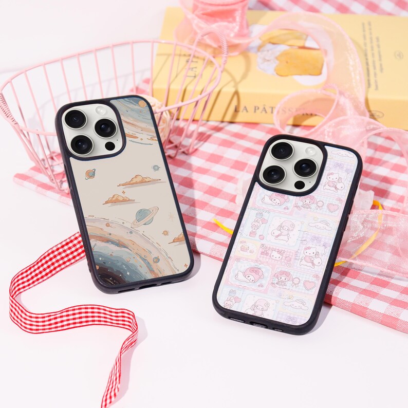 Op de afbeelding: Twee telefoonhoesjes met zwarte randen. E&eacute;n hoesje heeft een ruimte-thema ontwerp met planeten en wolken. De andere heeft een roze en wit patroon met stripfiguren. Beide hoesjes liggen op een roze en wit geruit oppervlak.