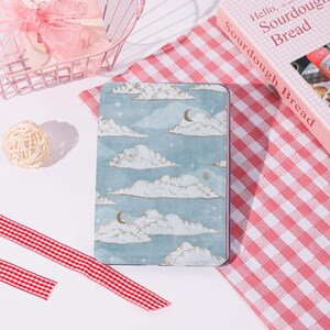 Peut inclure: Un carnet rectangulaire avec un motif de ciel et de nuages. La couverture présente un fond bleu clair avec des nuages blancs, des croissants de lune et des étoiles. Un tissu à carreaux roses et blancs et un livre intitulé "Hello, Sourdough Bread" sont en arrière-plan.