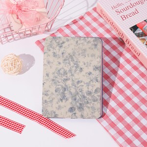 May include: A notebook featuring a blue and white floral pattern. It rests on a red and white gingham cloth, with a pink wire basket and a ball of twine nearby. A book titled "Hello, Sourdough Bread" is also visible in the background.