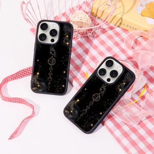 Puede incluir: Dos fundas negras para teléfono con un diseño celestial. Las fundas presentan una ilustración dorada de planetas y estrellas sobre un fondo oscuro. Las fundas tienen un borde oscuro y se muestran sobre una superficie a cuadros rosa y blanco.