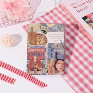 Puede incluir: Una caja metálica decorativa con ilustraciones de la película Ratatouille, con el texto "Anyone Can Cook!". También se ve un libro titulado "Hello, Sourdough Bread" y un mantel a cuadros rosa.