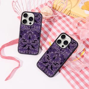 May include: Two phone cases with a purple and black bat pattern. The cases have a black border and a design featuring bats and ornate swirls. The cases are on a pink and white checkered surface.