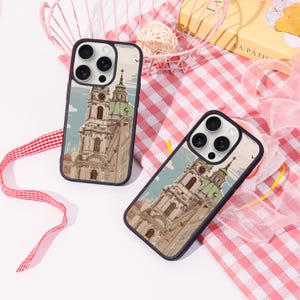 Peut inclure: Deux coques de téléphone avec une illustration détaillée d'une église européenne avec un dôme vert et un clocher. Les coques ont un bord bleu foncé et sont posées sur un fond à carreaux roses et blancs.