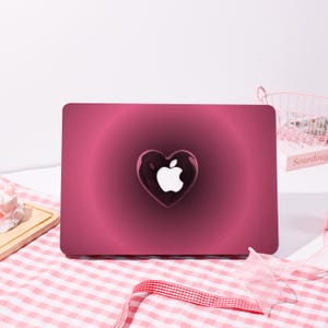 May include: A magenta laptop skin with a heart-shaped design and the Apple logo. The heart is a glossy, transparent dark red, and the Apple logo is white. The background has a radial gradient effect.
