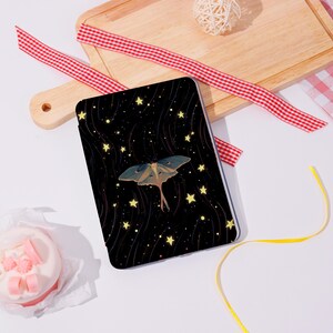 May include: A black tablet case with a celestial design. It features a moth with blue and green wings, and yellow stars against a dark background. The case is rectangular and likely made of a durable material.