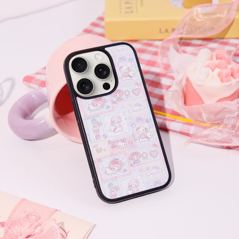 Op de afbeelding: Een zwarte telefoonhoes met een kleurrijk patroon met stripfiguren. De hoes heeft een zwarte rand en een camera-uitsparing. De achtergrond omvat een roze mok, een gebakje en een rood-wit geruite doek.