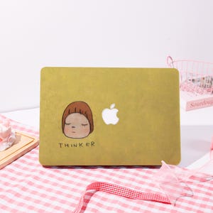 May include: A mustard-yellow laptop case with a cartoon illustration of a person with closed eyes and the word "THINKER" below. The Apple logo is visible on the right side of the case. The case is on a white surface with a pink and white checkered cloth.