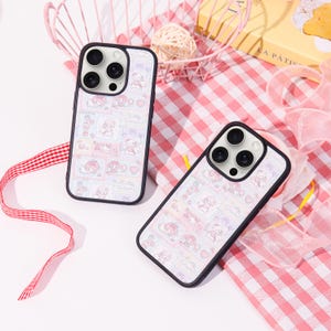 May include: Two phone cases featuring a colourful pattern of cartoon characters, hearts, and clouds. The cases have a black border and a white background. They are displayed on a pink and white checked surface.