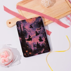 Pode incluir: Uma capa retangular para tablet com uma cena de Halloween. O design apresenta silhuetas de bruxas voando em vassouras contra um céu roxo e laranja, com um castelo assombrado e abóboras.