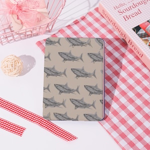 Könnte beinhalten: Ein rechteckiges Notizbuch mit einem sich wiederholenden Muster aus grauen Hai-Illustrationen auf beigem Hintergrund. Das Notizbuch liegt auf einem rot-weiß karierten Stoff. Ein Buch mit dem Titel "Hello, Sourdough Bread" ist sichtbar.