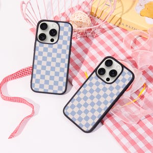 May include: Two phone cases with a blue and white checkered pattern. The cases have a black border and a camera lens cutout. The cases are on a pink and white checkered surface.
