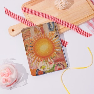 May include: A rectangular notebook with a colorful sun design on the cover. The sun is yellow and orange, surrounded by a collage of vintage-style labels and illustrations. A red and white checkered ribbon and a wooden cutting board are in the background.
