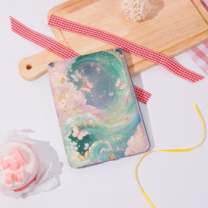 Può includere: Una custodia per tablet con un design stravagante con nuvole pastello, farfalle e dettagli dorati. La custodia ha un bordo trasparente e un'estetica sognante e artistica. La custodia è rettangolare.