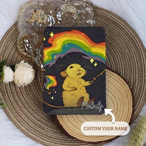 Puede incluir: Un cuaderno con una ilustración de dibujos animados de una rata amarilla creando un arcoíris con una varita mágica. El cuaderno tiene el nombre "Melody" y las palabras "CUSTOM YOUR NAME". El fondo es gris oscuro.
