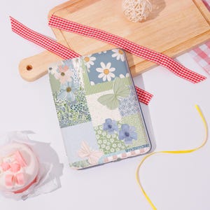 Pode incluir: Uma capa para tablet com um design floral em patchwork com margaridas, borboletas e flores azuis. A capa tem um esquema de cores azul claro, verde e branco. Uma fita xadrez vermelha e branca e uma tábua de corte de madeira estão no fundo.