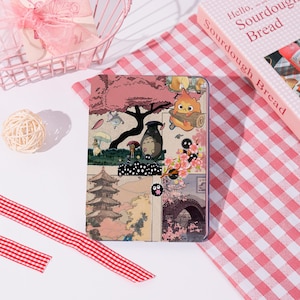 May include: A rectangular case with a collage of Japanese anime-style illustrations. The design includes characters, cherry blossoms, and traditional Japanese architecture. The case is on a pink and white checkered surface.
