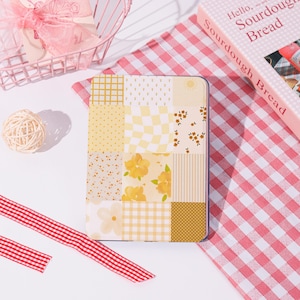 Könnte beinhalten: Eine rechteckige E-Reader-Hülle mit einem Patchwork-Design aus gelben und weißen Mustern, darunter Karos, Blumen, Punkte und Streifen. Die Hülle zeigt ein Sonnensymbol und verschiedene Blumenillustrationen in Gelb- und Cremetönen.