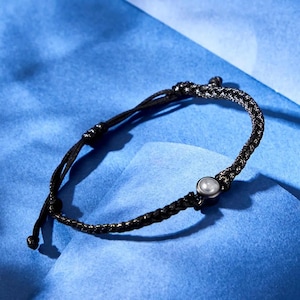 May include: A black and silver braided bracelet with a small, round, silver-colored bead. The bracelet is on a blue surface. The bracelet is adjustable.