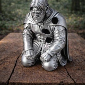 May include: A silver-colored figurine of a kneeling person with long hair, wearing a belted tunic and a cape. The figure features three distinct circular holes on the chest and shoulder area, with a somber facial expression and clenched fists.