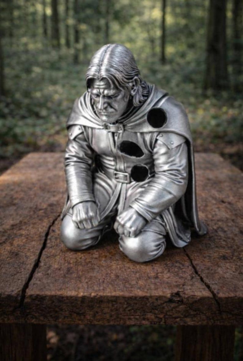May include: A silver-colored figurine of a kneeling person with long hair, wearing a belted tunic and a cape. The figure features three distinct circular holes on the chest and shoulder area, with a somber facial expression and clenched fists.