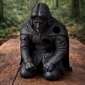 May include: A matte black sculpture of a kneeling figure with long hair, wearing a cloak and tunic. The figure has a somber expression with hands clenched in front of the knees. Several circular voids are visible on the chest and shoulder area.