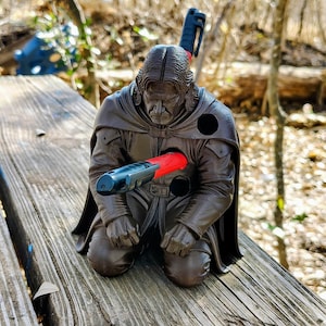 Könnte beinhalten: Braune Figur einer knienden Figur mit schwarzem Umhang und langem Haar, durchbohrt von einem rot-schwarzen Objekt. Die Figur befindet sich auf einer Holzoberfläche mit verschwommenem natürlichem Hintergrund.