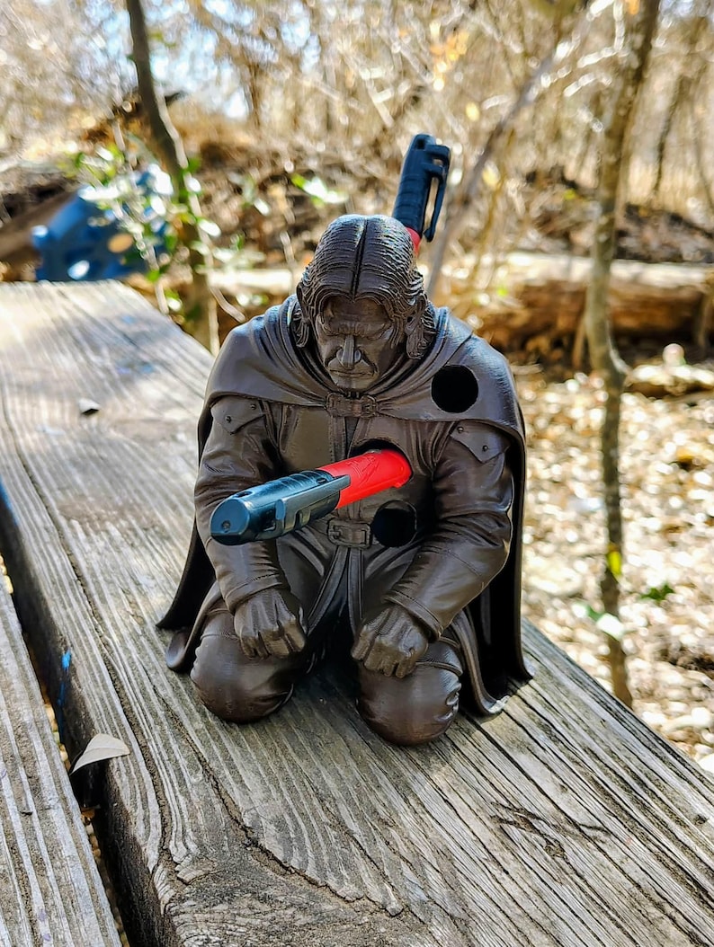 Op de afbeelding: Bruine beeldje van een knielende figuur met een zwarte cape en lang haar, doorboord door een rood en zwart object. De figuur staat op een houten oppervlak, met een vage natuurlijke achtergrond.