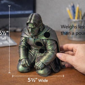 Op de afbeelding: Een knielend beeldje van een man in een mantel, ongeveer 16,5 cm hoog en 14 cm breed. De figuur heeft een metallic groene en zilveren afwerking. De tekst in de afbeelding luidt: "Weegt minder dan een pond."