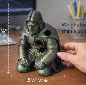 May include: A kneeling figure sculpture with a metallic green and bronze finish, featuring a hooded cloak and three circular cutouts. The figure is 6.5 inches tall, 5.5 inches wide, and weighs less than one pound, as indicated by the text.