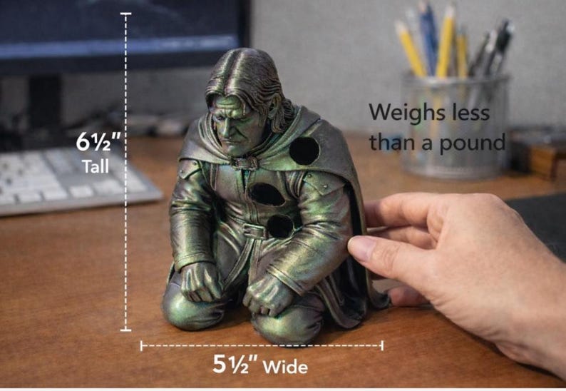 May include: A kneeling figure sculpture with a metallic green and bronze finish, featuring a hooded cloak and three circular cutouts. The figure is 6.5 inches tall, 5.5 inches wide, and weighs less than one pound, as indicated by the text.
