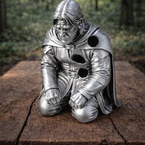 Op de afbeelding: Zilverkleurig beeld van een knielende figuur met lang haar en een cape. Het beeld staat op een houten oppervlak, met een bosachtergrond. De figuur draagt een tuniek met drie cirkelvormige uitsparingen.