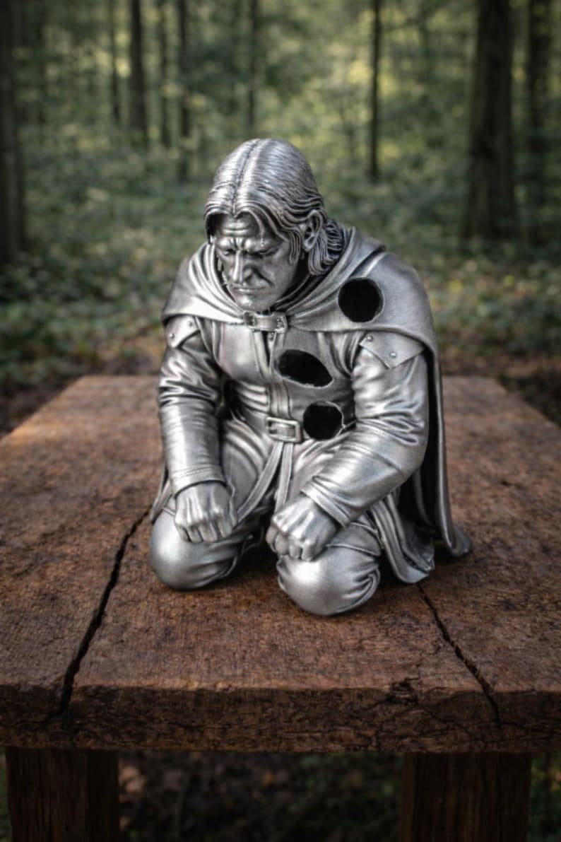 Op de afbeelding: Zilverkleurig beeld van een knielende figuur met lang haar en een cape. Het beeld staat op een houten oppervlak, met een bosachtergrond. De figuur draagt een tuniek met drie cirkelvormige uitsparingen.