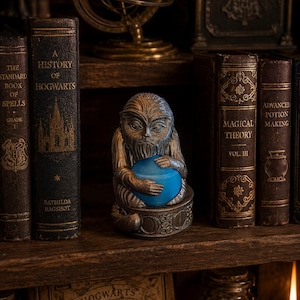 May include: A bronze-toned figurine of a seated creature with a bearded face and pointed ears, holding a smooth, bright blue sphere. The figure rests on a circular base featuring embossed circular patterns, positioned between two antique-style books.